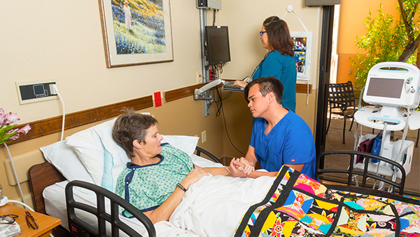 Healthcare professional in blue scrubs holds hands with a patient lying in a hospital bed with a colorful quilt, another healthcare worker uses a computer in the background.